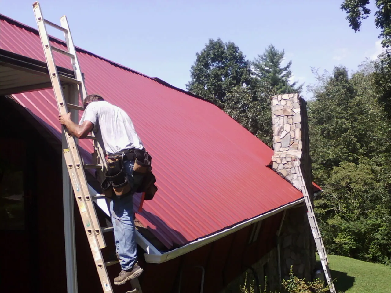 Roofer on a ladder beside a red metal roof and stone chimney — Soffit & Fascia Repair by Roofing Brothers in Kemmerer